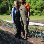A Black man and woman stand in a field of low growing vegetables wearing boots and farming overalls