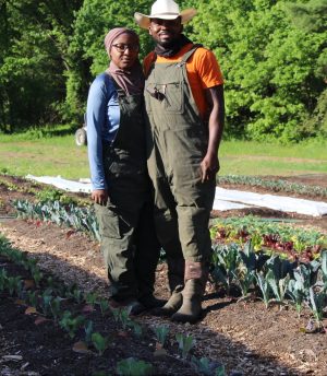 A Black man and woman stand in a field of low growing vegetables wearing boots and farming overalls
