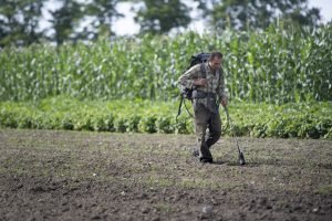a man walks through a bare field using a flame weeder worn on his back, with plants growing behind