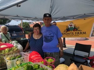 Man an women in  a side embrace standing behind table of produce underneath popup tent