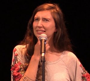 A woman in a pink shirt stands in front of a microphone with hand on chin and puzzled look on face