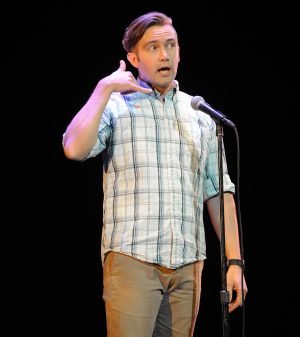 Man in plaid shirt on stage in front of microphone mimes speaking into phone