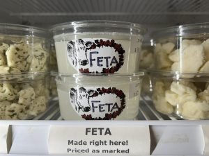 small plastic tubs of feta cheese (off-white chunks) and cheese curds (light yellow lumps) lined up on white wire racks in a fridge