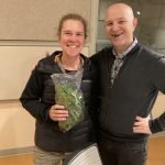 woman with bushy light brown hair in dark clothing stands next to bald headed man in dark clothing. Woman holds bag of spinach. Man has large piece of spinach comically in his teeth. Both are grinning