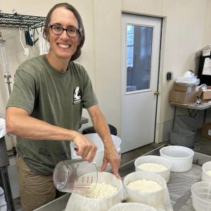 Woman wearing greens smiles at camera, pours curds and whey into round white plastic molds lined with mesh. stainless steel kitchen equipment in background